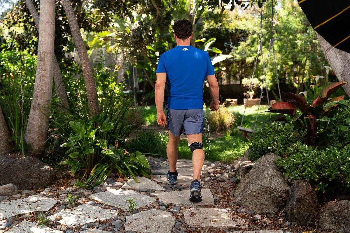 Athletic man walking thorough nature away from the camera while PEMF device is strapped around his knee.